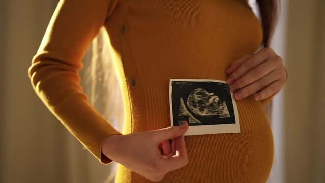 Pregnant woman looking ultrasound report. Happy pregnant female watching her ultrasound report and touching her abdomen, admiring sonography picture of her baby. Expecting baby, pregnancy concept.