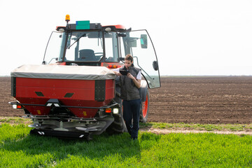 Young farm worker next to tractor using digital tablet. Tractor spreading artificial fertilizers in...