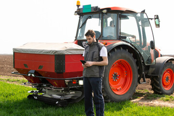 Young farm worker next to tractor using digital tablet. Tractor spreading artificial fertilizers in...