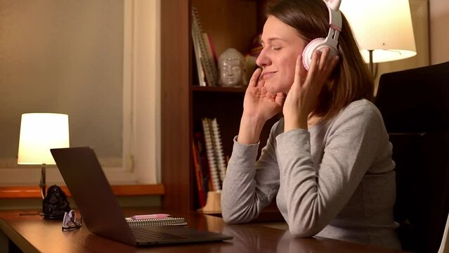A 30s woman in her home office listening to music on headphones. She is having a break, relaxing, and enjoying herself, smiling and moving her head to the rhythm of the music, happy and relaxed mood.
