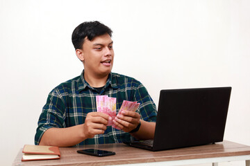 Young asian college student sitting counting Indonesia banknotes while using laptop on the desk...