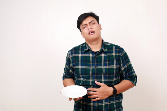 Young asian man feels hungry, touching his stomach while holding an empty plate