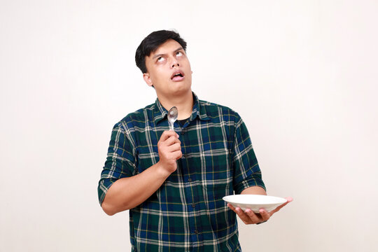 Thoughtful young asian man holding an empty plate while imaging something