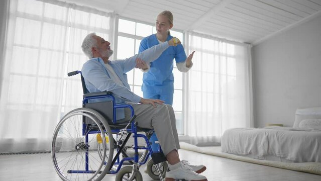 Man in wheelchair exercising with nurse. Social medical worker woman helping pensioner doing physical hands exercises with dumbbell. Palliative care in late stage of neurodegenerative pathologies.