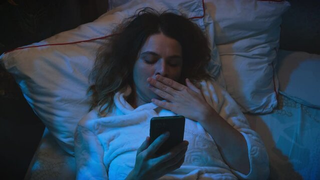 Insomnia concept. Sleepless tired young woman lying in bed scrolling smartphone at night. Unhealthy lifestyle.