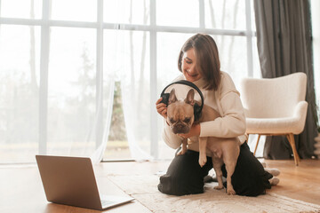 Wearing headphones. Young woman is with her pug dog at home