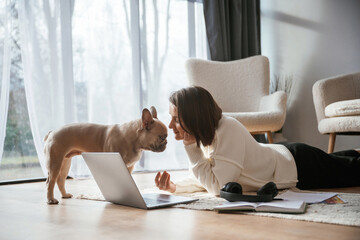With laptop. On the floor. Young woman is with her pug dog at home