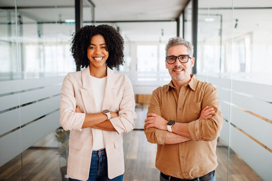 A radiant businesswoman and her mature male colleague stand with arms crossed in a modern office, exuding confidence and professional equality. Diverse work team concept