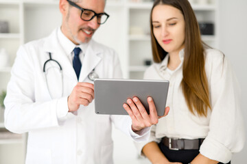A mature doctor is engaging with a young female patient, showing her something on a digital tablet,...