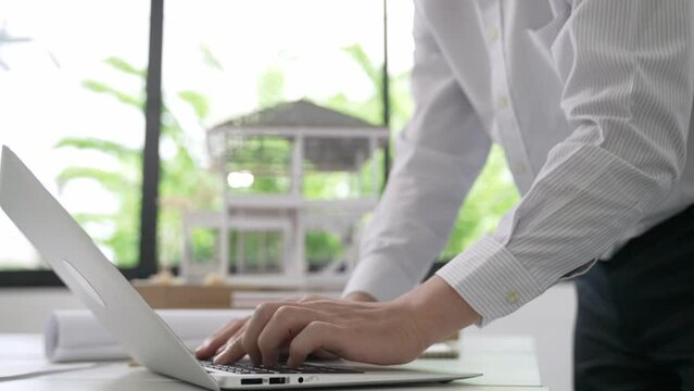 Close up of professional male architect engineer measure house model and using laptop analysis data at meeting table with house model and map . scatter around. Focus on hand. Immaculate.