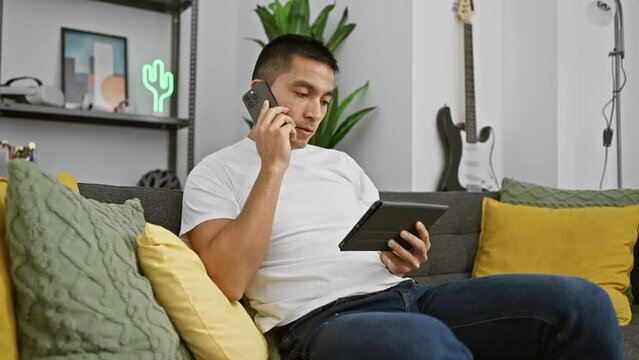 A hispanic man multitasks with a phone call and tablet in a cozy living room setting, exuding a casual, modern lifestyle.