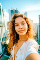 Young Latina woman in her 20s, with curly hair, taking a selfie on the rooftop overlooking a big city. Travel concept.