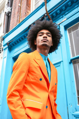 Latin man in his 30's with afro hair wearing an elegant orange suit next to a blue facade. High angle shot.