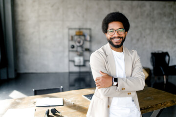 A cheerful Brazilian businessman in a smart casual beige suit stands with arms crossed in loft office, his smile reflecting confidence and approachability