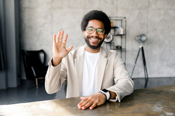 A cheerful young businessman greets with waving during virtual meeting, seated in a modern office...
