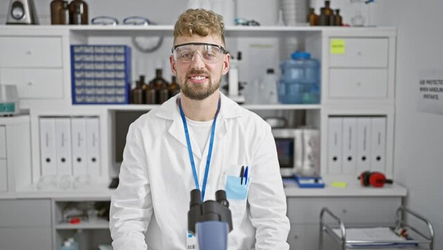 A Handsome Young Man With A Beard Wearing Safety Glasses And A Lab Coat Poses In A Well-equipped Research Laboratory.