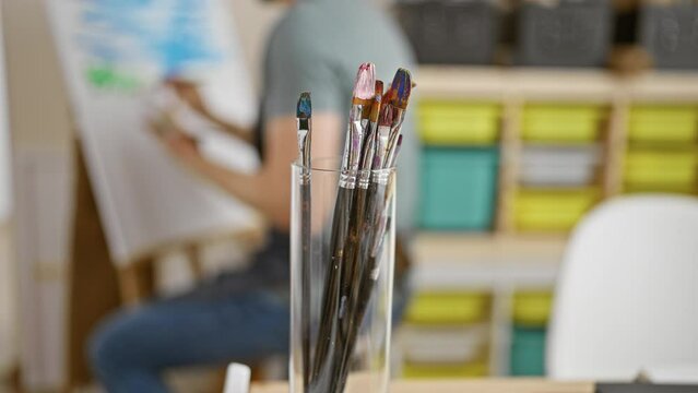 Close-up of paintbrushes in a glass with a blurred artist painting in a creative studio background