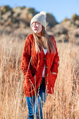 Young woman outdoors in tall grass wearing rust colored sweater and knit cap.