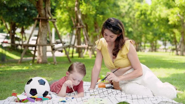 Beautiful Caucasian woman, young mother, sits near handsome son lying on small mat, where two people have picnic, mother child are doing activities together, child lies down paints with hands.