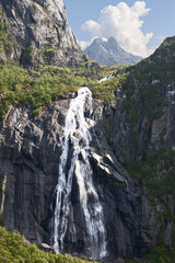 Valefossen waterfall flows vigorously down the cliffs of Lofoten Islands, Norway, amidst lush foliage and under a dynamic summer sky. - obrazy, fototapety, plakaty