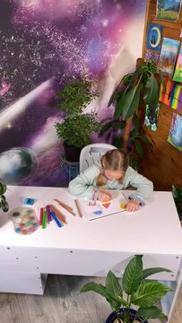 Youthful artistic imagination, childhood hobby. Vertical lovely little girl expresses her creativity by joyfully drawing with colored pencils surrounded by artistic supplies, pictures, indoor plants