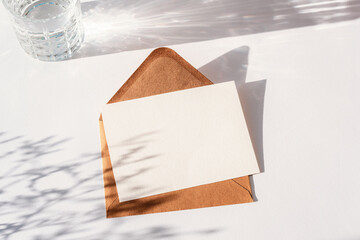 Envelope with blank card, glass of water and flowers shadow on white table. Top view, flat lay,...