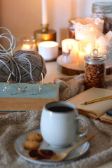 Cup of tea or coffee, cookies, books, glasses, e-reader, almonds, ball of yarn, knitting needles and lit candles on the table. Hygge at home. Selective focus.
