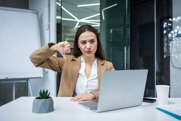 nervous woman manager showing thumb down and expressing negative emotions. Caucasian female feeling disappointment while working at laptop in personal designer cabinet. - obrazy, fototapety, plakaty