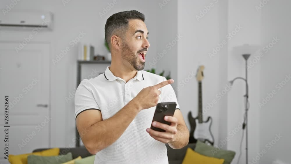 Poster cheerful young hispanic man joyfully pointing off to the side while texting and laughing at home. th