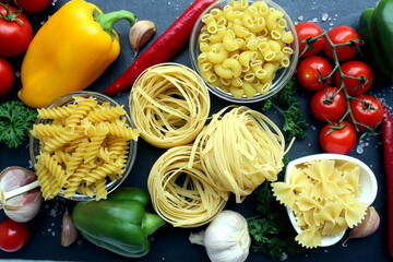 On a black stone board lie different pasta in plates, pasta nests and vegetables.