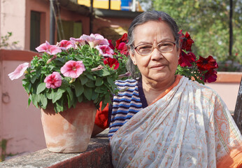 a simple dressed aged Bengali woman happily looking at potted winters flowering plants, namely pansy and petunias. The plants are looking beautiful with vibrant coloured blooms. In a rooftop garden du