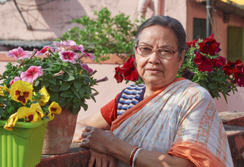 a simple dressed aged Bengali woman standing beside vibrant looking blooming winter flowers , namely pansies and petunias. Shot in a rooftop garden during winter in Kolkata.