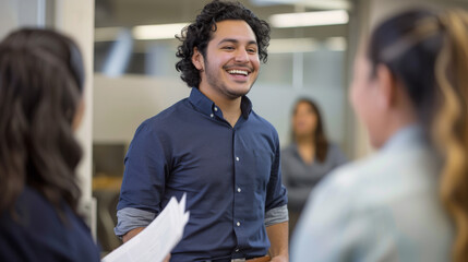 A man in a casual shirt is laughing and holding a document during a cheerful interaction with colleagues in an office setting. - obrazy, fototapety, plakaty