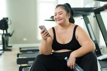 Overweight Asian woman exercise in gym, smiles while talking on phone, taking joyful break from her fitness routine. With a beaming smile, young fitness enthusiast enjoys a conversation on her phone