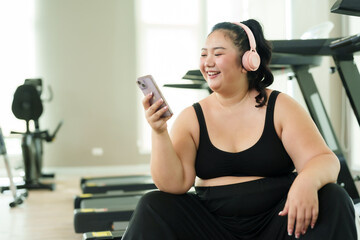 Fototapeta na wymiar Plus-size Asian woman exercise in gym, female in activewear joyfully uses her smartphone during break, reflecting balance of leisure and fitness. Sitting comfortably on a treadmill, happy engages