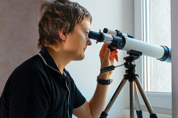 A man looks through a telescope from the room. Side view of a young man looking out the window and...