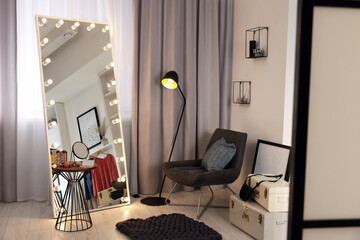 Makeup room. Stylish mirror with light bulbs, beauty products on table and armchair indoors