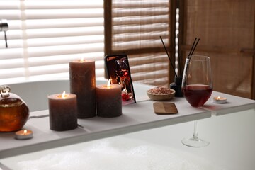 White wooden tray with smartphone, glass of wine and burning candles on bathtub in bathroom