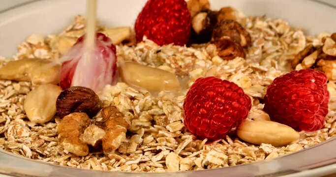 Macro video in which oat milk is poured into a bowl of spelled muesli with spelled flakes, raspberries, hatelnuts, walnuts and almonds.