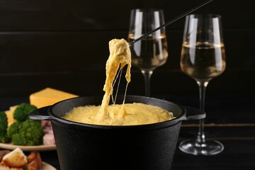 Dipping piece of ham into fondue pot with melted cheese on table, closeup