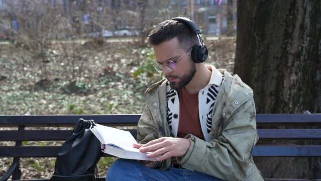 Young handsome hipster freelance business man wearing jacket reading a book while sitting outdoors. Businessperson on lunch break listening music on wireless headset and read novel to clear his mind