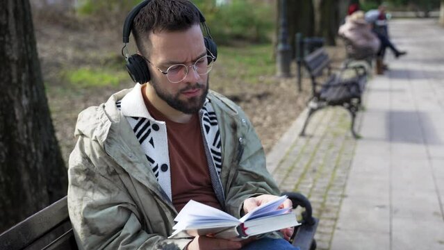 Young handsome hipster freelance business man wearing jacket reading a book while sitting outdoors. Businessperson on lunch break listening music on wireless headset and read novel to clear his mind