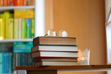 Stack of books, reading glasses and lit candles on the table. Rainbow bookshelf in the background. Selective focus.