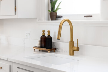 A gold kitchen faucet detail with white cabinets and soap dispensers sitting on a white marble...