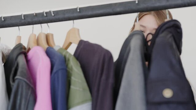 The image presents a young woman with a contemplative expression, browsing through a selection of garments hanging on a clothes rack. The focus is on her as she thoughtfully examines the textures and