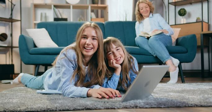 Beautiful smiling girls younger and eldest lying on the floor and use laptop computer while their loving carefree woman mother reading book on the couch near them in sitting room