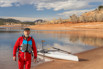 senior man wearing drysuit and life jacket with a rowing shell on lake shore