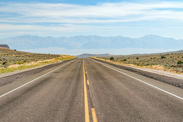 Fototapeta na wymiar Deserted asphalt highway through the Nevada desert summer landscape with haze covered mountain range and blue sky with clouds. USA.