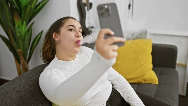 Hispanic woman vaping indoors while using her smartphone, surrounded by home decor in a cozy apartment setting.