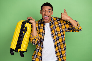 Photo of handsome positive man hold suitcase demonstrate thumb up approval isolated on green color background